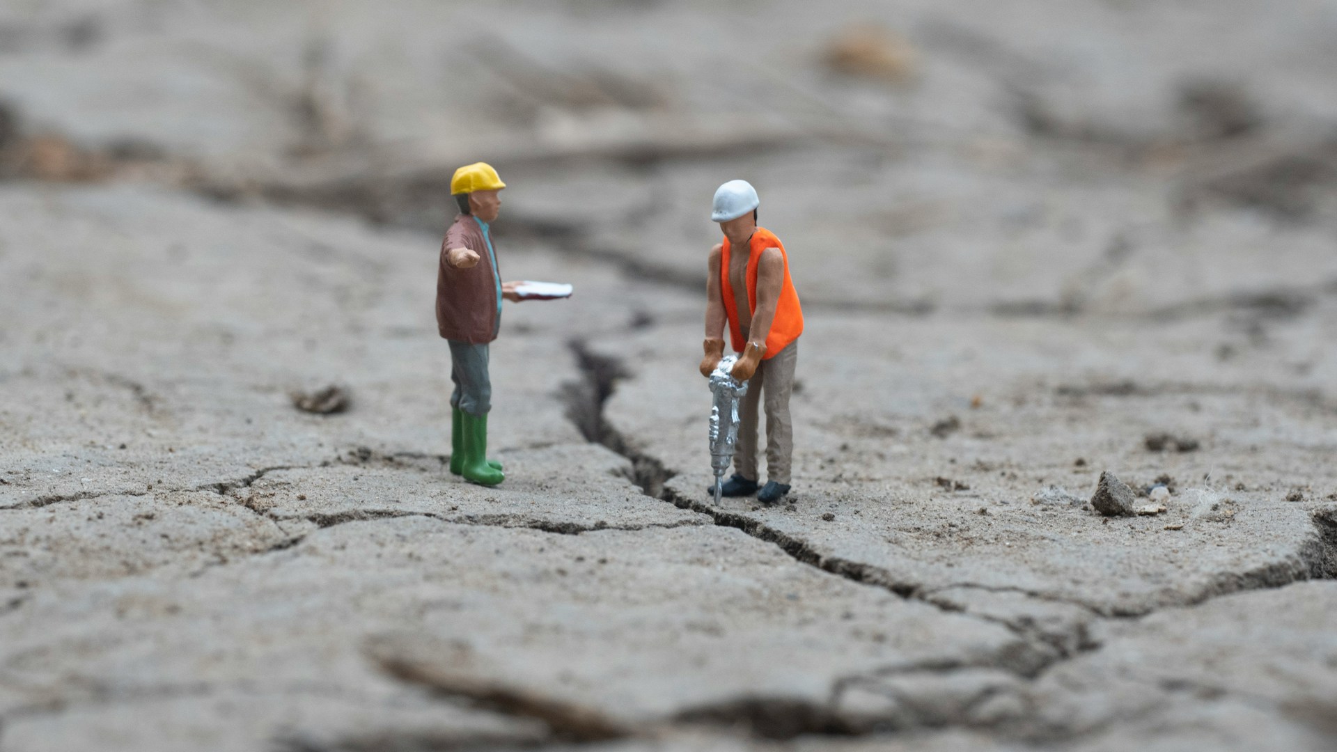 Two miniature construction worker figurines on cracked, dry earth - one with a jackhammer, one supervising with a clipboard - as fractures spread across the ground beneath them.