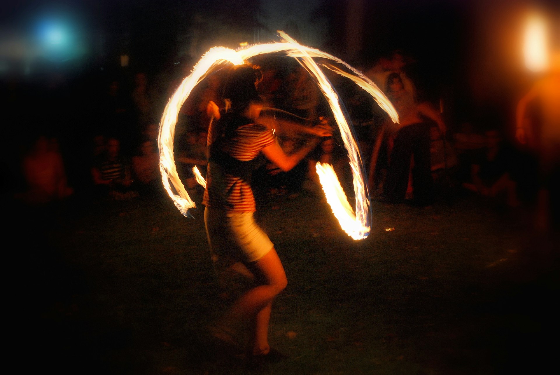 A figure mid-spin inside a self-made ring of fire, body blurred by long exposure, barefoot on dark grass while onlookers watch from the edges.