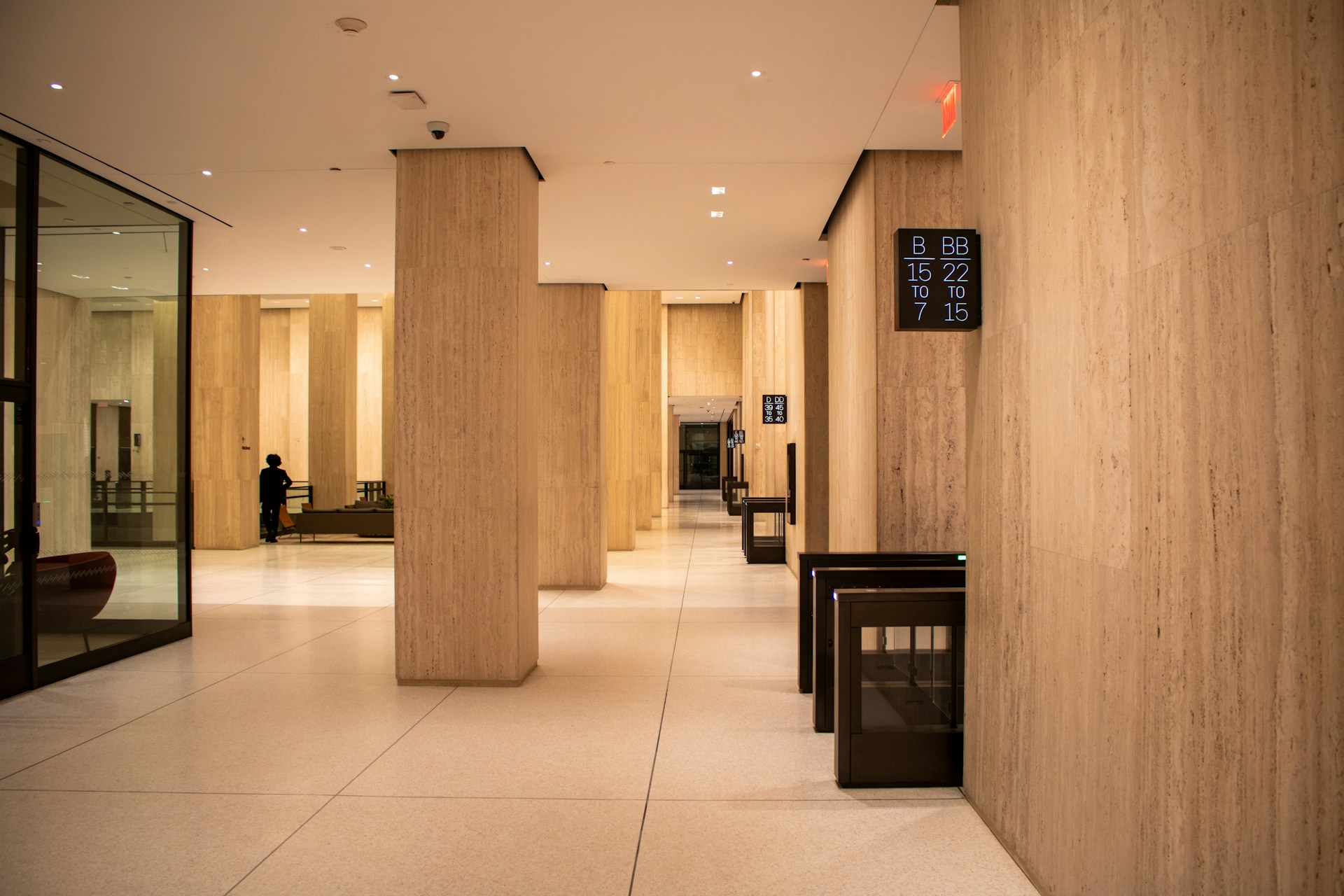 A cavernous travertine-clad corporate lobby, warmly lit, with stone columns, empty access turnstiles, and a single figure standing alone in the middle distance.