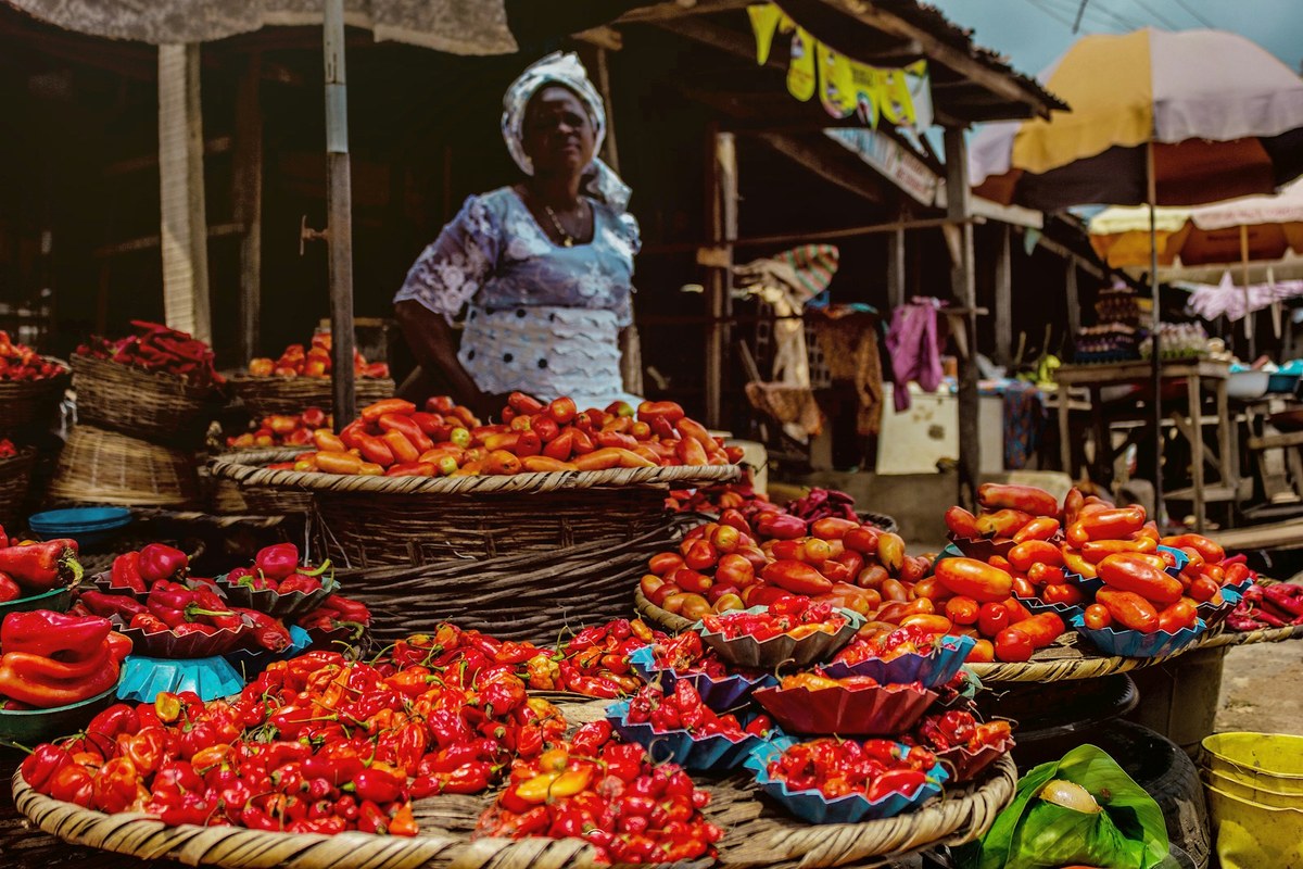A woman in white lace and a head wrap stands behind baskets of red peppers and tomatoes at an outdoor market.